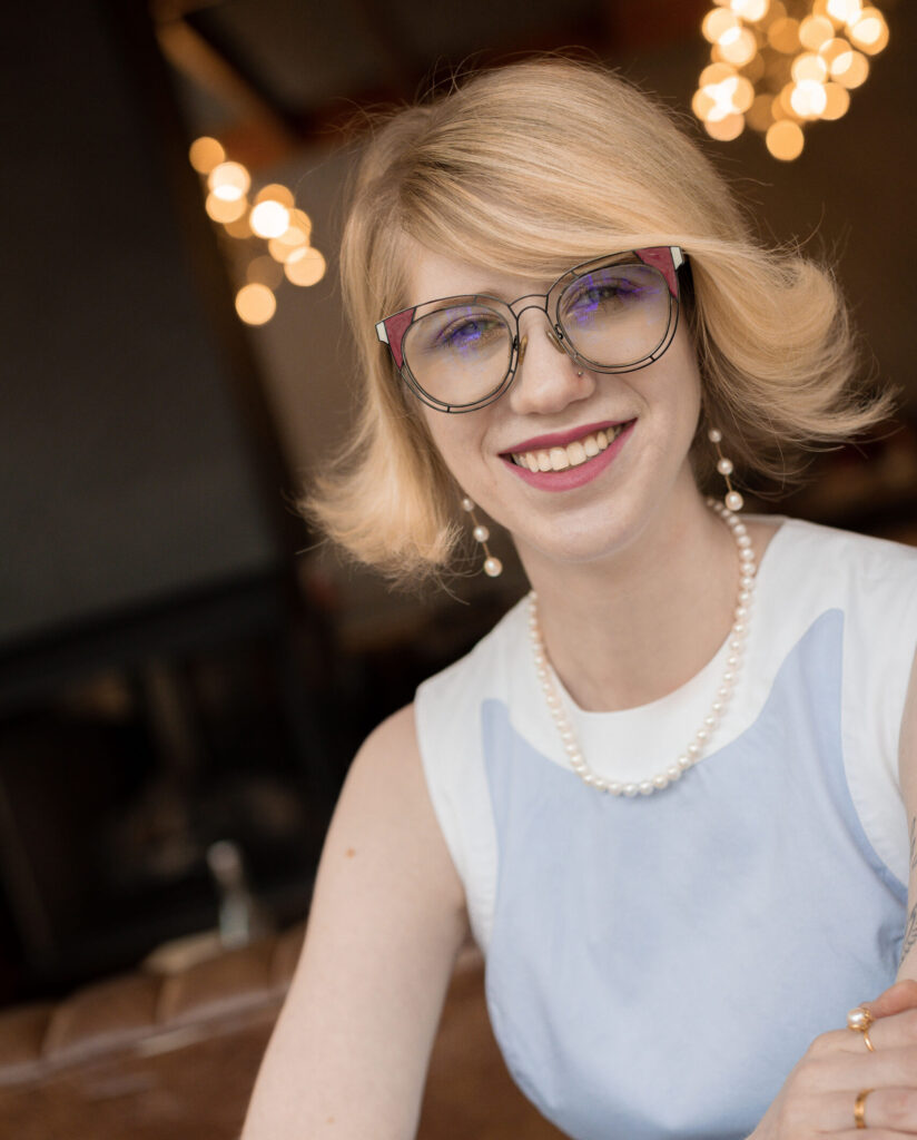 photo of cassandra, a smiling woman with short blonde hair wearing large glasses with black frames. she has pink lipstick on and is wearing a pearl necklace. she is dressed in a light blue and white colorblocked sleeveless dress as she leans on a table with her arms crossed. in the background, there are blurred lights giving a warm bokeh effect.